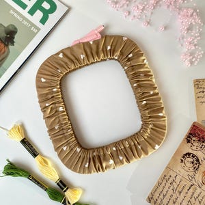 May include: A square, ruched, tan fabric frame with white heart patterns. A pink clamp is attached to the frame. Spools of yellow and green thread are next to the frame. A magazine and pink beaded garland are also in the image.