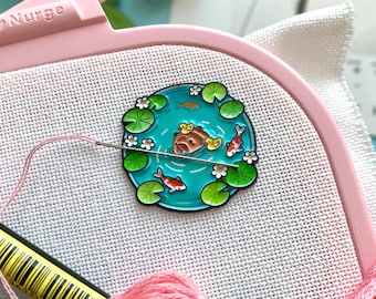 May include: A cross-stitch project in a pink embroidery hoop. The project features a colorful pond scene with a bear, fish, and lily pads. Pink and green embroidery floss and a needle are also visible.