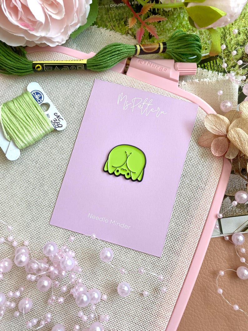 Op de afbeelding: Een limoengroene emaille naaldhouder in de vorm van een achterwerk, op een roze kaart met "Ms Pattern" en "Needle Minder". De naaldhouder ligt op een canvas oppervlak met groen borduurgaren en roze bloemaccenten.