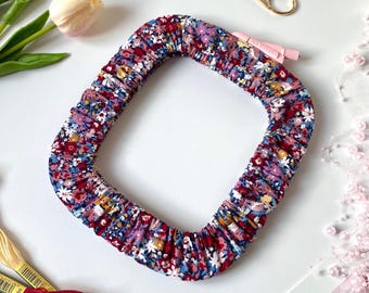 May include: A floral patterned, square-shaped hair accessory in shades of red, blue, and white. The scrunchie is surrounded by other craft supplies, including embroidery floss, scissors, and flowers.