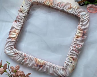 Puede incluir: Un aro de bordado rosa y blanco con un estampado floral y de pájaros. El aro está sobre una superficie blanca con un carrete de hilo de bordar y un protector de agujas.