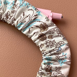 May include: A floral fabric hair accessory with a pink plastic applicator. The fabric features a cream background with brown and blue floral patterns. The accessory is curved, suggesting it is a headband or similar item.