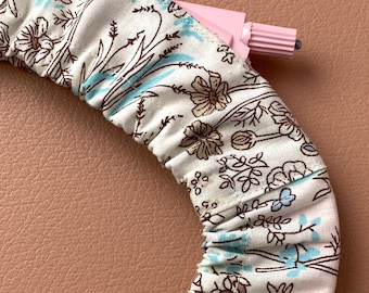 Puede incluir: Un accesorio para el cabello de tela floral con un aplicador de plástico rosa. La tela presenta un fondo crema con estampados florales marrones y azules. El accesorio es curvo, lo que sugiere una diadema o artículo similar.