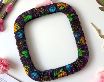May include: A square embroidery hoop cover featuring a black fabric base with a vibrant, multicolored mushroom and star pattern. The cover has an elasticized edge for a snug fit and includes a light pink plastic tension adjustment knob at the top.