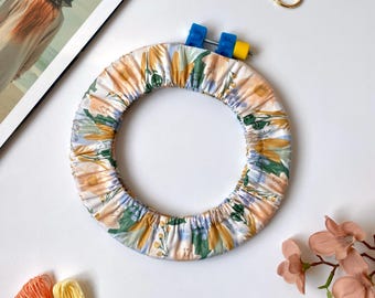Puede incluir: Un bastidor floral con un cierre azul y amarillo, tijeras doradas, hilo de bordar y una revista. El bastidor presenta un patrón de flores amarillas, naranjas y verdes sobre un fondo blanco. La revista dice "ER" y "SPRING 2017 $50."