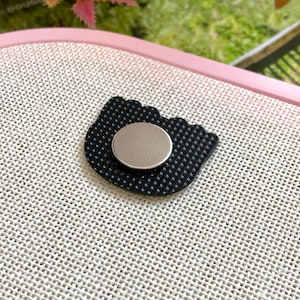 Op de afbeelding: Een zwarte, katvormige bedel met een getextureerd oppervlak en een zilverkleurige, cirkelvormige magneet aan de achterkant. De bedel ligt op een lichtgekleurde, getextureerde achtergrond.