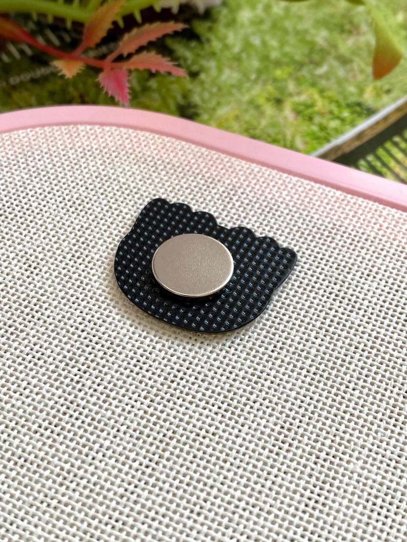 Op de afbeelding: Een zwarte, katvormige bedel met een getextureerd oppervlak en een zilverkleurige, cirkelvormige magneet aan de achterkant. De bedel ligt op een lichtgekleurde, getextureerde achtergrond.