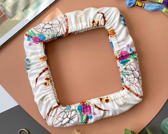 May include: A white, square-shaped pillowcase with a floral and dreamcatcher design. The pillowcase has a colorful pattern with pink, blue, and green flowers. The background is a mix of brown, gray, and white.