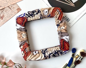 May include: A square, patterned fabric frame with a floral and geometric design. The frame is in shades of brown, red, and blue. The image also includes embroidery floss in blue, cream, and brown, along with a magazine and postcards.