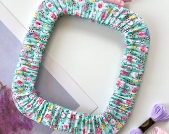 May include: A rectangular embroidery hoop cover featuring a light mint green fabric with a floral pattern of pink, white, and yellow blossoms. The cover is gathered with elastic and includes a small pink plastic tension adjustment knob.