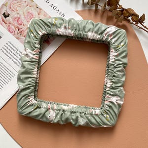 Op de afbeelding: Een groene stoffen ring met een wit konijn en bloemenpatroon. De ring ligt op een lichtbruin oppervlak.