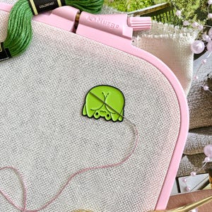 Op de afbeelding: Een roze borduurring houdt witte stof vast met een groen en gele emaille pin. Groen borduurgaren en een gouden schaar zijn ook zichtbaar. De ring is gemarkeerd met de merknaam "AD Nurge".