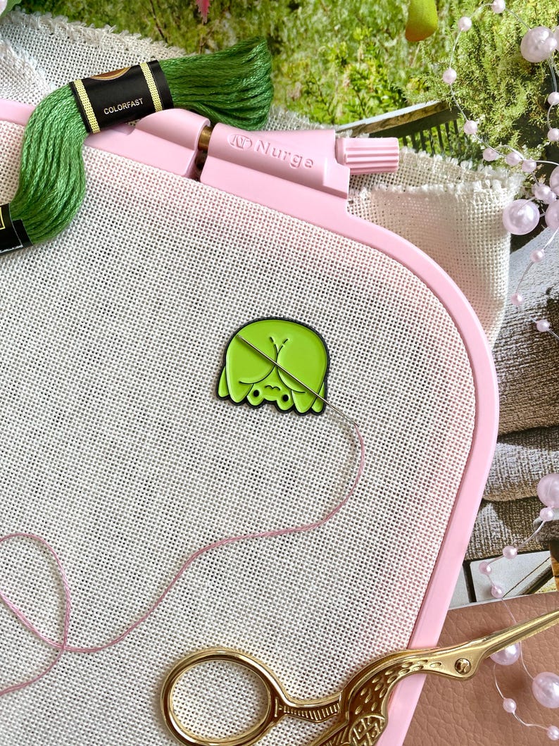 Op de afbeelding: Een roze borduurring houdt witte stof vast met een groen en gele emaille pin. Groen borduurgaren en een gouden schaar zijn ook zichtbaar. De ring is gemarkeerd met de merknaam "AD Nurge".