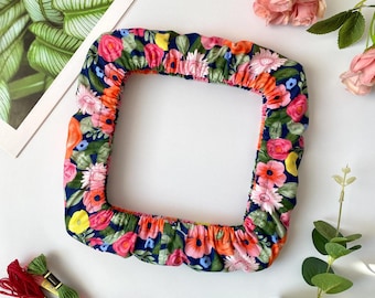 May include: A square, floral-patterned fabric item with a navy blue background and a colorful array of pink, orange, and yellow flowers. The fabric is gathered around the edges. Embroidery floss in red, green, and pink is also visible.