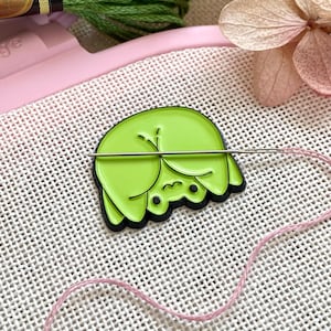 Op de afbeelding: Een limoengroene emaille pin in de vorm van een cartoon kikker met zwarte accenten, doorboord door een naald en roze draad, rustend op een witte kruissteek stof. Groen borduurgaren en een roze borduurring zijn op de achtergrond.