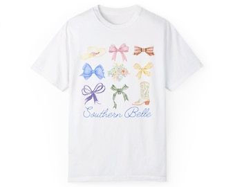 May include: White t-shirt featuring a watercolor design of bows, a hat, flowers, and a cowboy boot. The words "Southern Belle" are written in a cursive font below the images. The shirt is made of a soft, comfortable material.