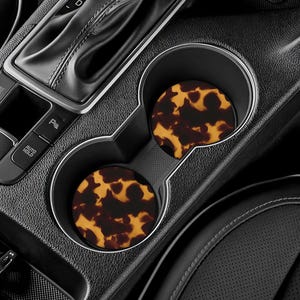 May include: Two car coasters with a tortoiseshell pattern, placed in a car's cup holders. The coasters have a brown and yellow design, and the car interior is black.