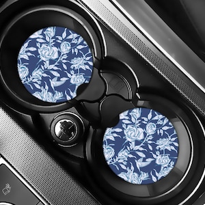 May include: Two car coasters with a blue floral pattern featuring white roses and leaves. The coasters are circular and sit inside a black car console cup holder. The design is a repeating pattern of roses and foliage.