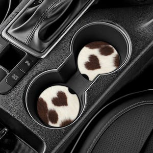 May include: Two car coasters with a cowhide pattern, featuring brown and white spots, are placed in a car's cup holders. The coasters are round and add a decorative touch to the vehicle's interior.