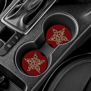 May include: Two car coasters with a red background and a leopard print star design. The coasters are placed in a car's cup holders, surrounded by black interior components.