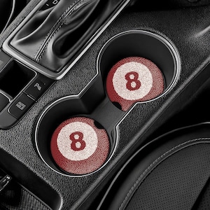 May include: Two car coasters with a red and white 8-ball design are placed in a car's cup holders. The coasters have a textured surface and a white number 8 in the center. The car's interior is black.