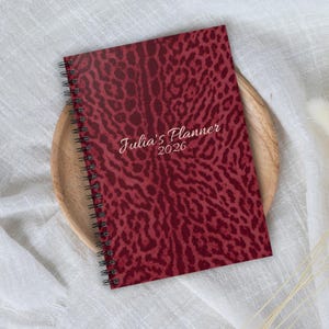 Può includere: Un'agenda rilegata a spirale con copertina con stampa leopardata bordeaux. Il testo "Julia's Planner 2026" è stampato in corsivo. L'agenda è appoggiata su un vassoio di legno e tessuto bianco.