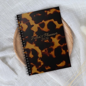 Puede incluir: Una agenda con espiral y cubierta con estampado de carey. La portada presenta el texto "Julia's Planner 2026" en letras doradas. La agenda está sobre una bandeja de madera, sobre un fondo de tela blanca.
