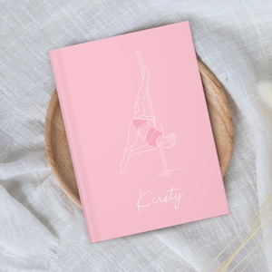 May include: A light pink notebook featuring a white line art illustration of a person performing a pole dance move. The cover includes the name Kirsty written in a white cursive script font below the central graphic design element.