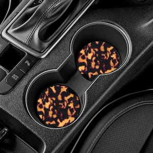 May include: Two car coasters with a tortoiseshell pattern sit in a car's cup holders. The coasters have a brown and yellow design. The car's interior is black, with a gear shift and other controls visible.