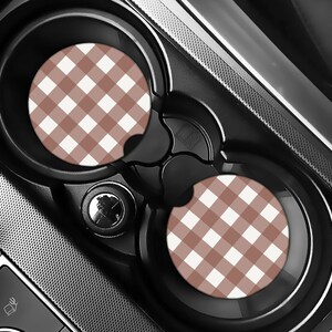 May include: Car cup holder coasters with a brown and white gingham pattern. The coasters are round and fit inside the car's cup holders. The car's interior is black and gray.