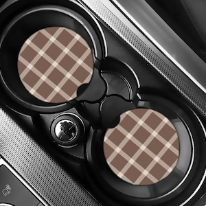 May include: Two car coasters with a brown and cream plaid pattern sit in a car's cup holders. The coasters are round and made of absorbent material. The car's interior is black and gray.