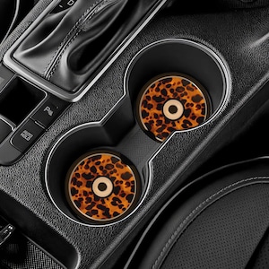 Op de afbeelding: Twee auto-onderzetters met een schildpadpatroon, met bruine en zwarte vlekken, liggen in de bekerhouders van een auto. De onderzetters hebben een lichtbruine cirkel in het midden. Het interieur van de auto is zwart.