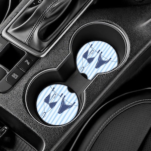 May include: Two car coasters with a blue and white striped background. Each coaster features a navy blue bikini hanging on a clothesline. The coasters are placed in a car's cup holders.