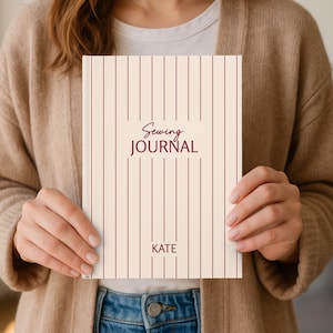 May include: A cream-coloured sewing journal with vertical burgundy stripes. The words "Sewing Journal" are printed in a script font, with the name "KATE" below. The journal is held by a person.