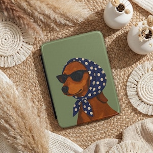 May include: An olive green tablet case featuring a brown dog wearing black sunglasses and a blue polka dot scarf. The dog illustration is centered on the case, which is surrounded by a woven texture.