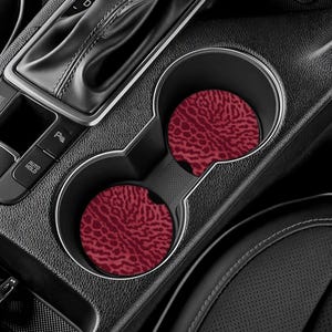 May include: Two car coasters with a burgundy animal print pattern are placed in a car's center console cup holders. The coasters are round and made of absorbent material. The car's interior is black.