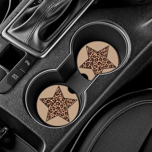 May include: Two car coasters with a tan background and a leopard print star design. The coasters are placed in the car's cup holders, with the car's gear shift and console visible in the background. The coasters add a decorative touch to the car's interior.