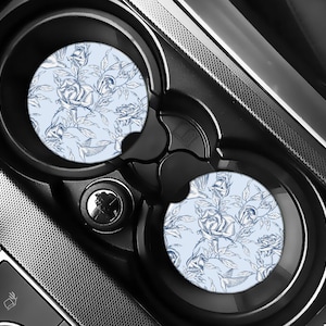 May include: Two car coasters with a floral rose design. The coasters feature a light blue background with a pattern of blue roses and leaves. The coasters are in a car's cup holders, surrounded by black plastic.