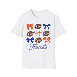 Könnte beinhalten: Weißes T-Shirt mit einem Design aus American-Football-Bällen und Schleifen in Rot, Blau und Orange. Das Wort "Florida" ist in einer Schreibschrift am unteren Rand des Designs geschrieben.