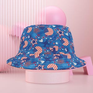 Könnte beinhalten: Ein blauer Fischerhut mit einem patriotischen Muster mit amerikanischen Flaggen, Sternen und anderen patriotischen Symbolen. Der Hut wird auf einem rosa Sockel vor einem rosa Hintergrund präsentiert. Der Hutrand ist mit rosa Stoff gefüttert.