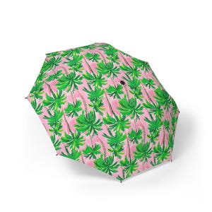 May include: A pink umbrella featuring a repeating pattern of green palm trees. The umbrella is open, displaying the bright colours and tropical design. The palm trees have green fronds and brown trunks.