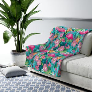 Puede incluir: Una manta vibrante con un diseño tropical de flamencos rosas, flores de hibisco y follaje verde sobre un fondo turquesa. La manta está sobre un sofá blanco, creando un acento colorido. El diseño evoca el verano y las vacaciones. Medidas: aproximadamente 150 cm x 200 cm.