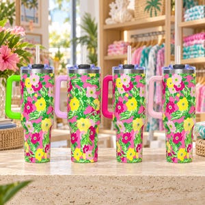 Könnte beinhalten: Vier Edelstahlbecher mit Griffen und transparenten Strohhalmen, jeweils mit einem lebendigen Blumenmuster aus rosa, gelben und grünen Blüten auf leuchtend rosa Hintergrund. Jeder Becher hat eine eigene Grifffarbe: Limettengrün, Lavendel, Pink und Hellrosa.