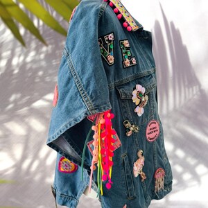 Könnte beinhalten: Hellblaue Jeansjacke mit bunten Aufnähern, floralen Applikationen und Perlenverzierungen. Die Jacke zeigt das Wort "VE" in floralen Aufnähern sowie rosa Pompons und bunte Quasten.