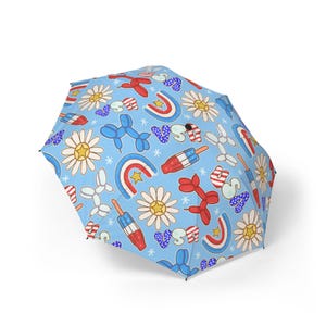 May include: A light blue umbrella featuring a repeating pattern of patriotic motifs in red, white, and blue. The design includes daisies, balloon animals, rainbows, and ice lollies. The umbrella is open and set against a white backdrop.
