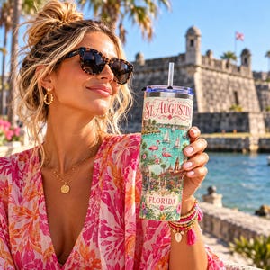 May include: A cylindrical stainless steel tumbler with a clear lid and straw, featuring a colourful graphic print of St. Augustine, Florida, including illustrations of sailboats, pink flamingos, and historic architecture with the text St. Augustine Florida.