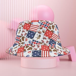 Könnte beinhalten: Ein Patchwork-Eimerhut mit roten, weißen und blauen Quadraten. Der Hut zeigt Bilder der amerikanischen Flagge, Sterne, Fußbälle, Traktoren und einen Adler. Der Hut wird auf einem rosa Sockel präsentiert.