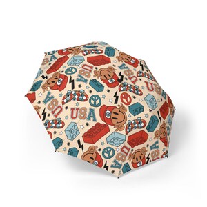 May include: A beige umbrella featuring a repeating pattern of red, blue, and tan graphics. The design includes smiling faces, building blocks, skateboards, peace signs, stars, and the word "USA".
