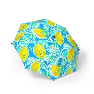May include: An open umbrella with a colourful pattern of yellow lemons, green leaves, and white flowers set against a bright blue backdrop. The umbrella has a black handle and a classic dome shape, ideal for protection from the elements.