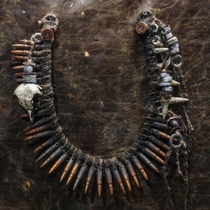 Puede incluir: Un collar hecho de cordón de cuero marrón, balas y otros abalorios de metal y hueso. El collar está colocado sobre una superficie de cuero marrón.